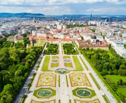 A wide view of formal gardens with symmetrical designs, fountains, and trimmed hedges leading to the palace.