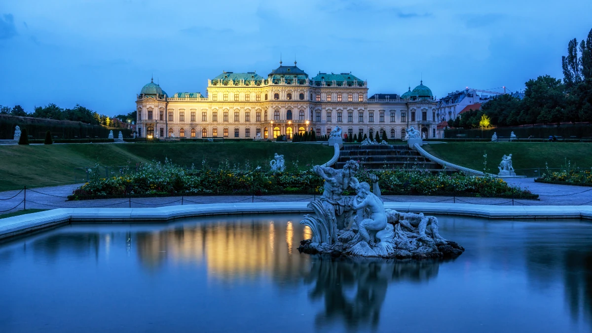 A night view of the Belvedere Palace in Vienna with its lights reflecting in the water and a statue fountain in the foreground.