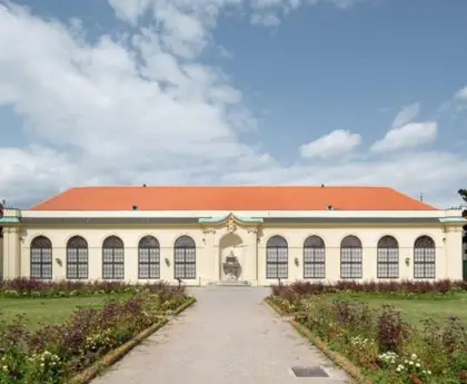 The Belvedere Orangery with white walls and a red roof, set against a partly cloudy sky and a garden path.