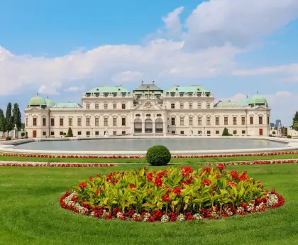 A grand baroque-style palace with a green copper roof and a flowerbed in front.