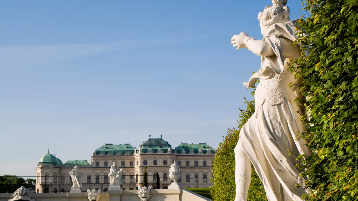  A white statue surrounded by greenery with Belvedere Palace visible in the distance.