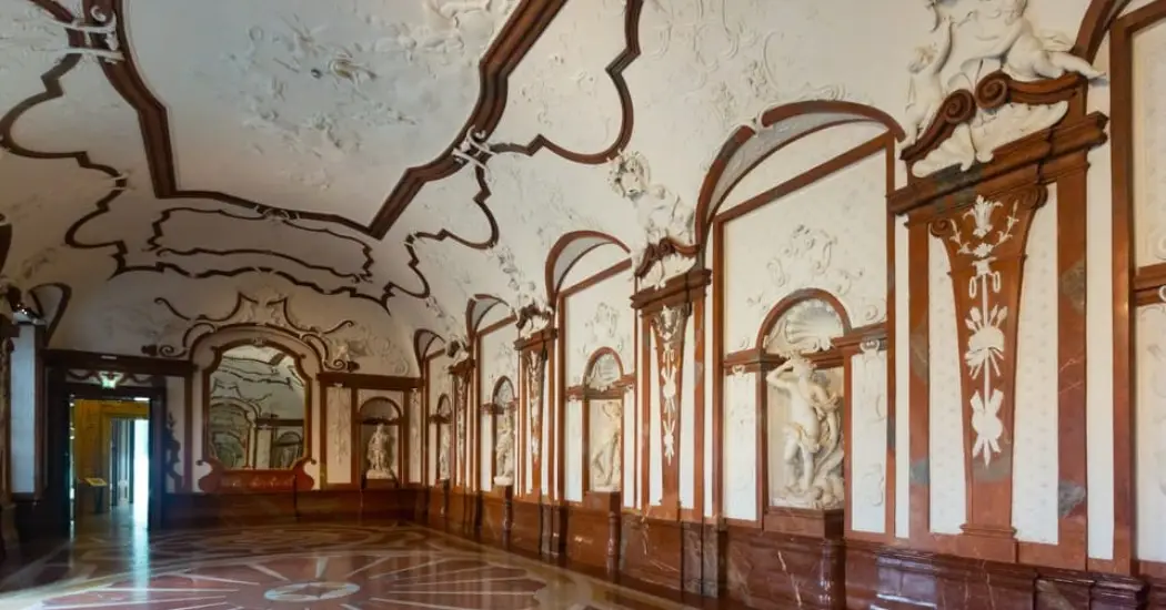 A long, elegant hallway in a palace with a detailed marble floor and white sculpted walls.