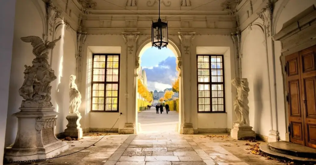 Interior passage of Lower Belvedere with sculptures and soft sunlight, leading to the gardens and the view of Upper Belvedere far away.