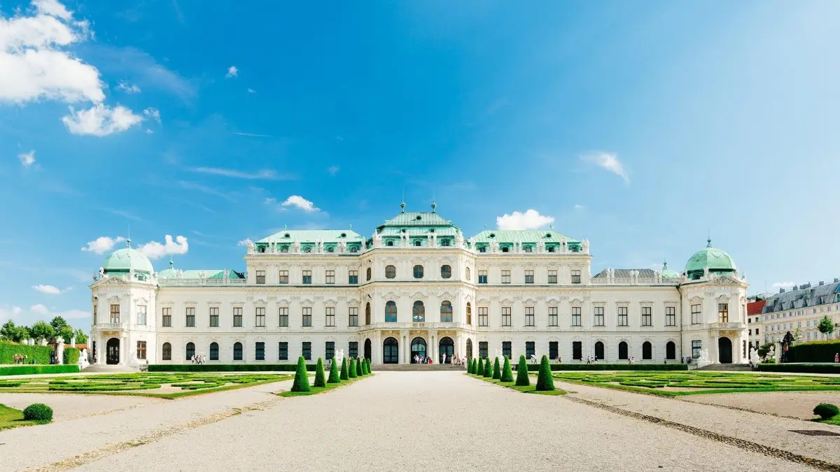 Front view of the Belvedere Palace in Vienna with manicured gardens, clear blue sky, and historic Baroque architecture featuring green domed roofs.