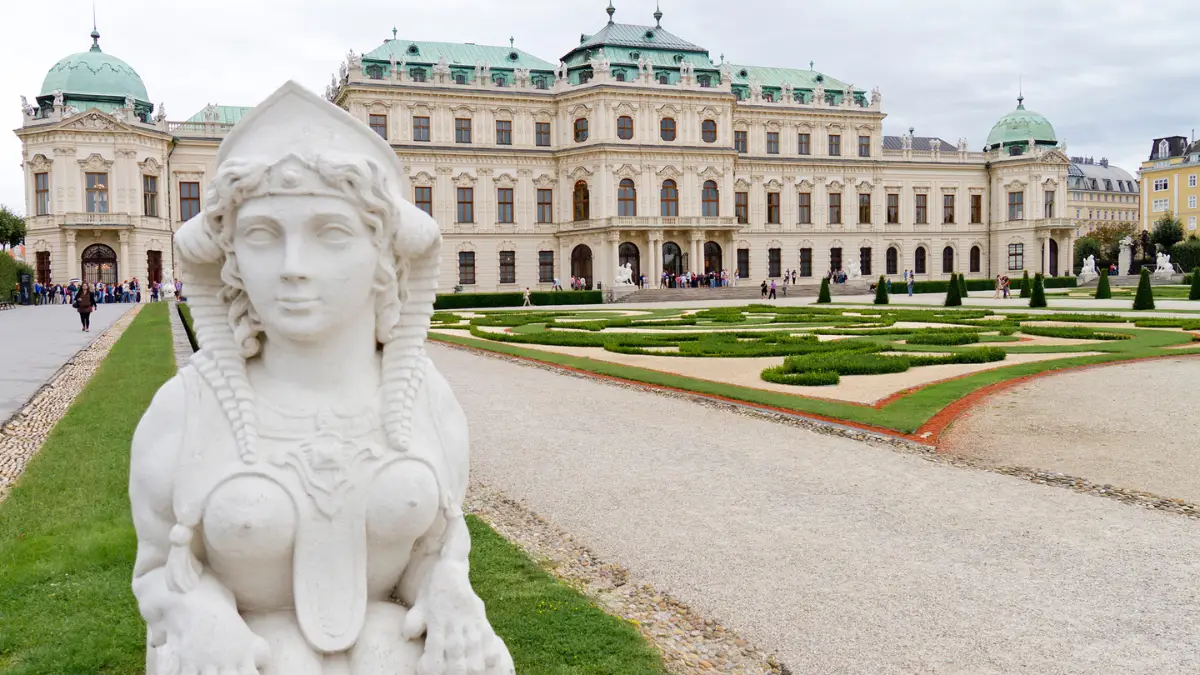 A white stone sphinx statue in a manicured garden with Belvedere Palace in the background.