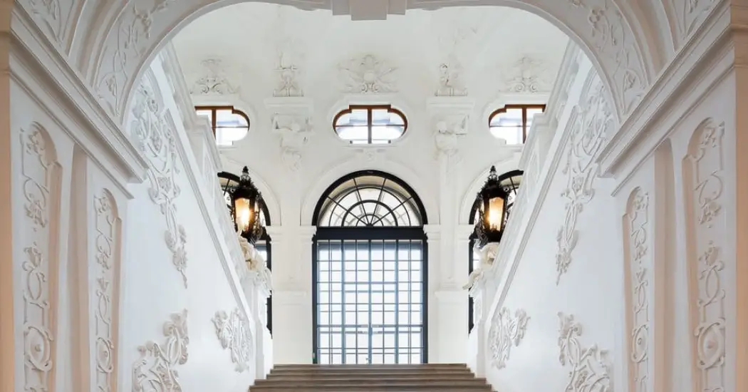 Ornate white staircase with intricate wall carvings and large arched windows inside Belvedere Palace, Vienna.