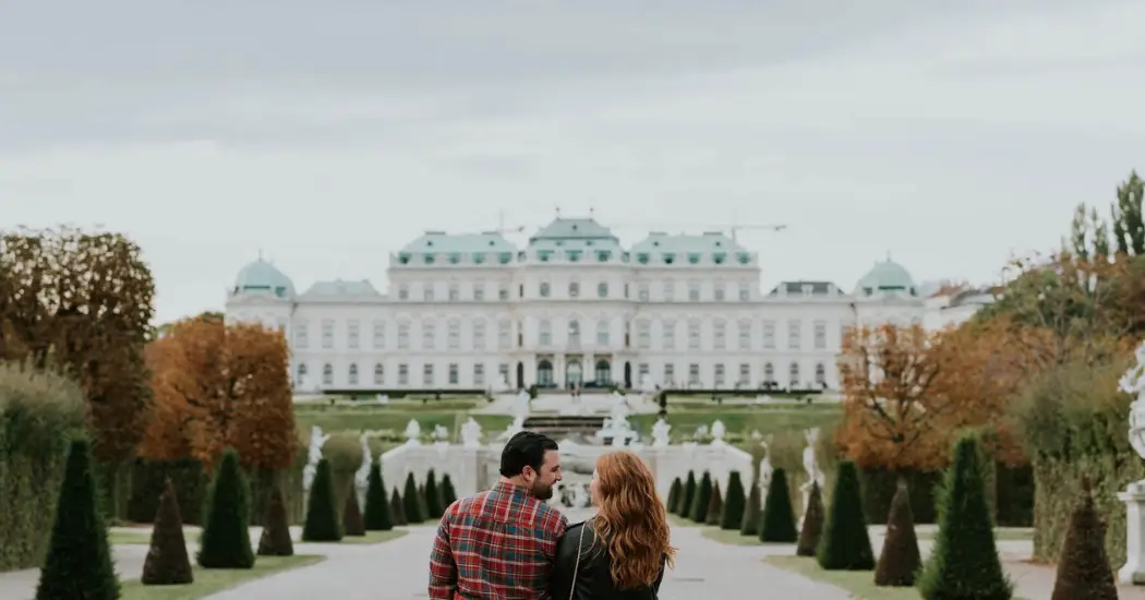 A couple walking hand-in-hand towards Belvedere Palace through manicured gardens with autumn trees lining the pathway.