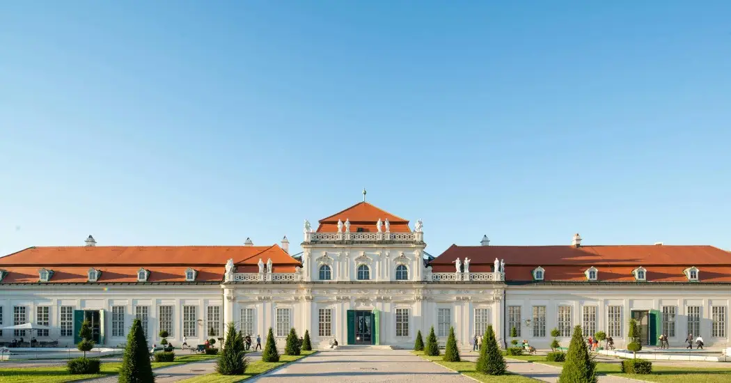 Lower Belvedere Palace in Vienna with red rooftops, white walls, statues on the roof, and a neat garden path lined with small cone-shaped trees. Lower Belvedere Palace in Vienna with red rooftops, white walls, statues on the roof, and a neat garden path lined with small cone-shaped trees.
