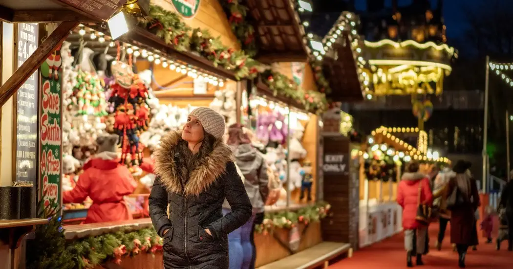 Tourists in winter attire admiring festive decorations at Christmas market with holiday stalls and a carousel in the background