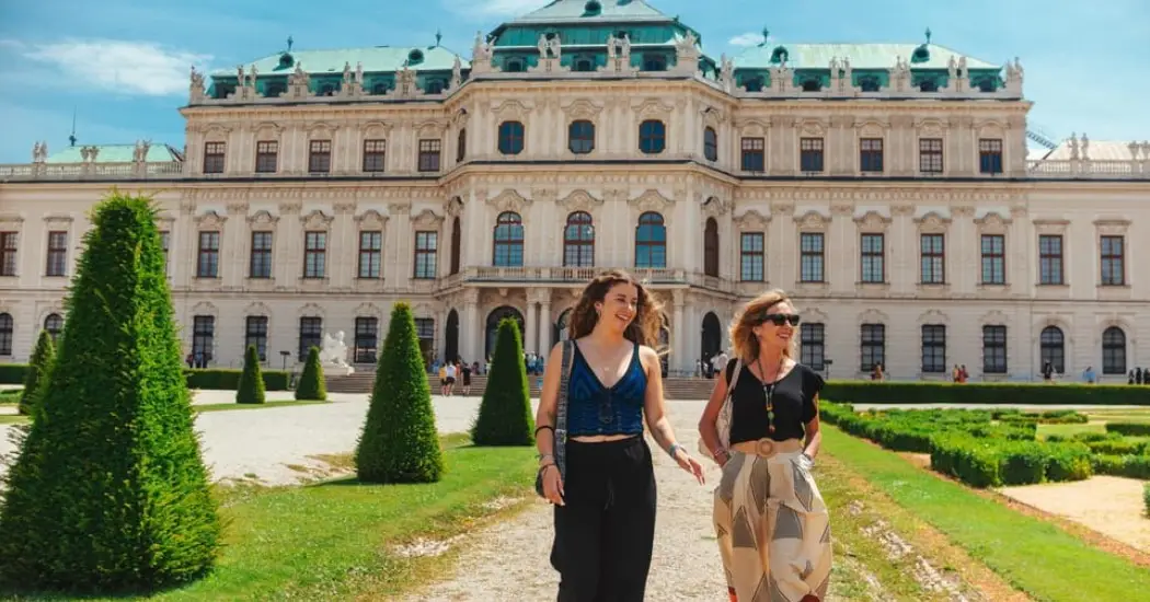 Tourists are walking and smiling in front of the Belvedere Palace in Vienna on a sunny day.