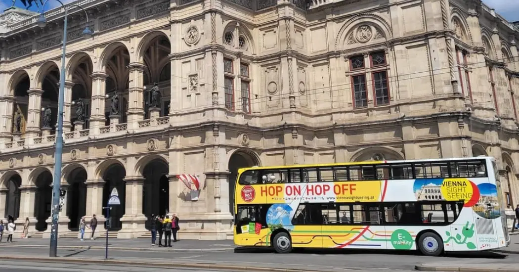 A yellow Vienna Sightseeing hop-on hop-off bus parked in front of the Vienna State Opera building.