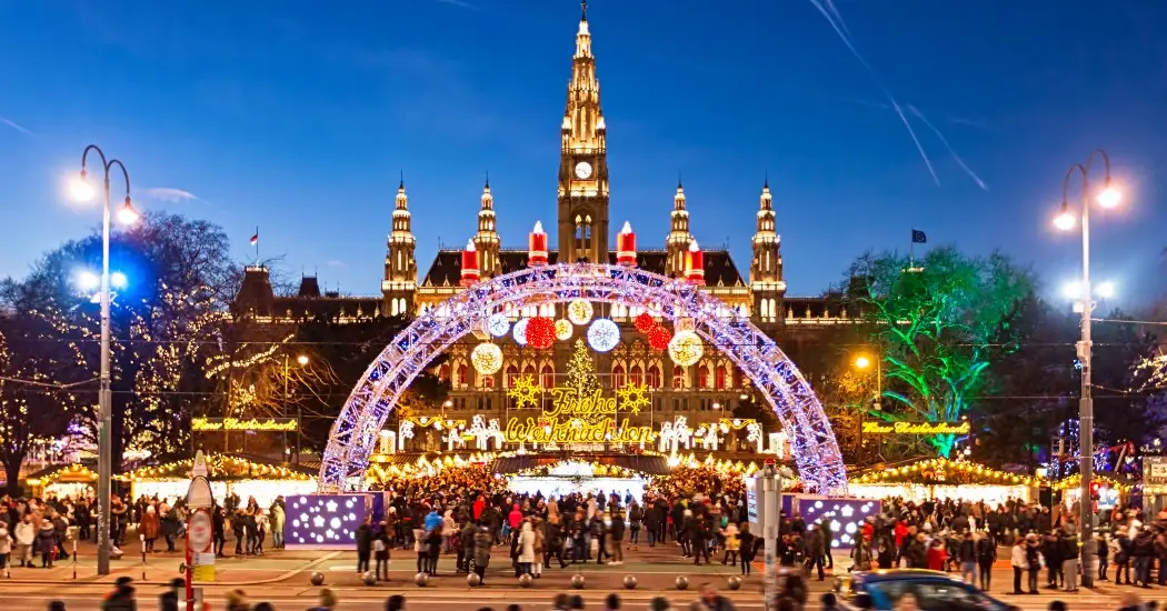 Festive crowds gathered at Vienna Rathaus Christmas Market under glowing arches and a lit-up city hall
